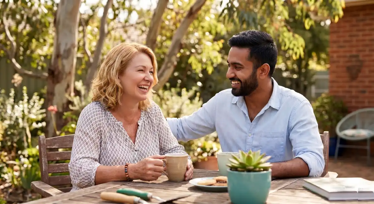 An older blonde woman and a young South Asian man sitting together at a wooden patio table outdoors, smiling warmly over coffee.